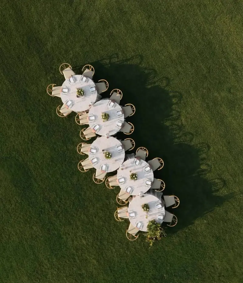 Overhead view of five round tables with white cloths and wood chairs in a row on a green lawn