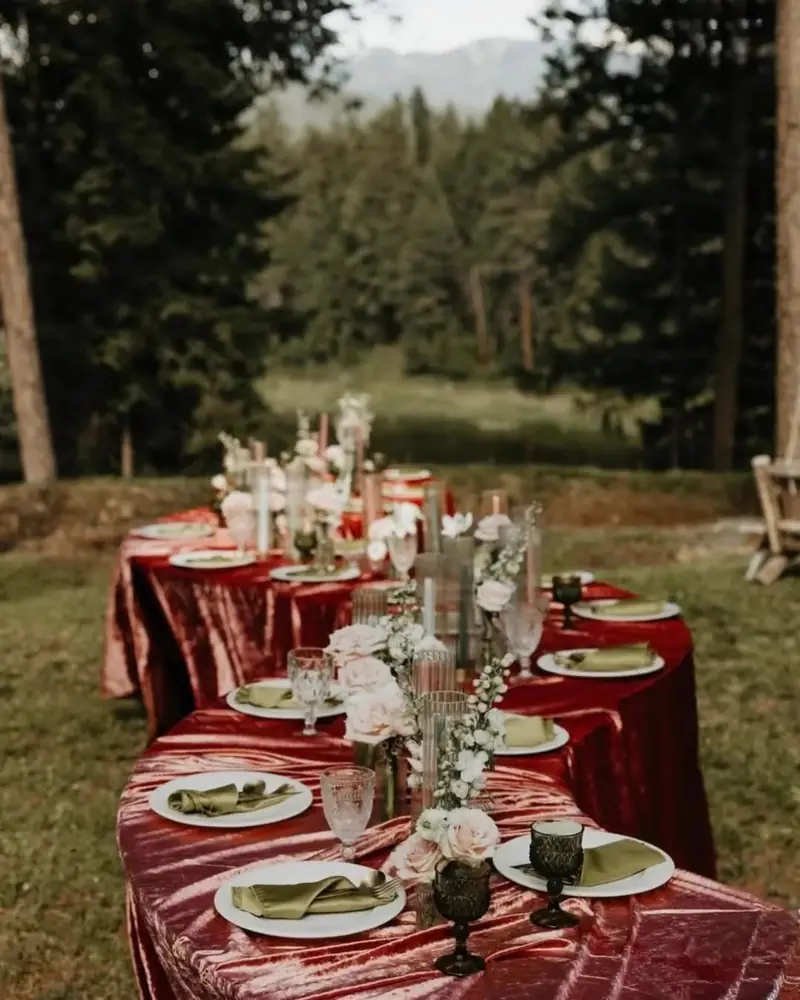 Long banquet table in a burgundy velvet cloth with green goblets and candles among pine trees