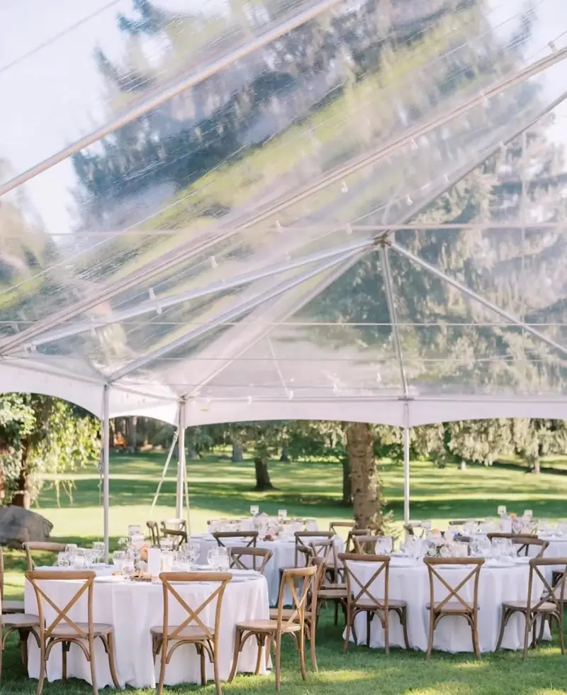 Round tables in white cloths with cross-back chairs and white flowers under a clear-top tent on a lawn