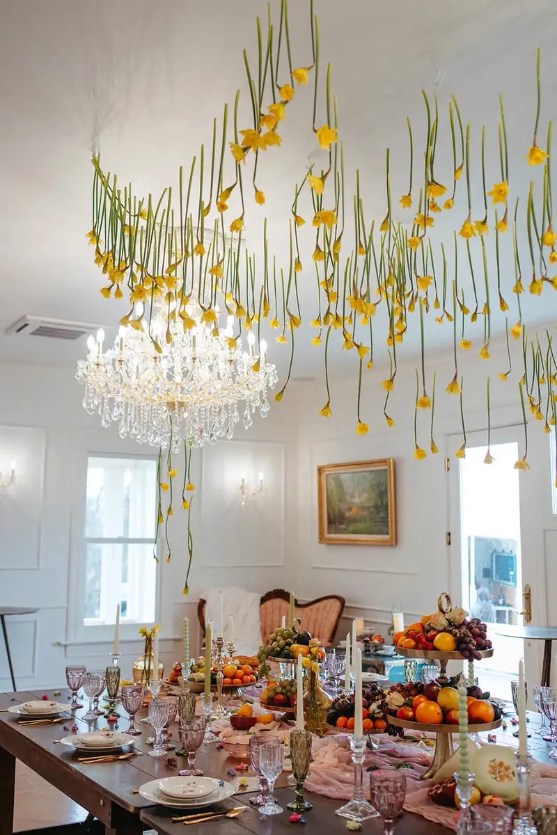 Yellow flowers hanging over a crystal chandelier above a long dining table with fruit stands