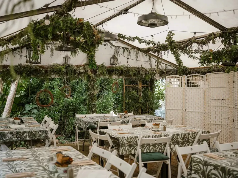 Tables in leaf-print cloths with white folding chairs under hanging greenery, lanterns and string lights
