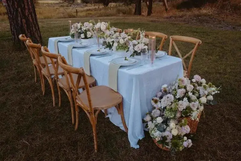 Long table in a light blue cloth with sage napkins and cross-back chairs beside a large tree outdoors