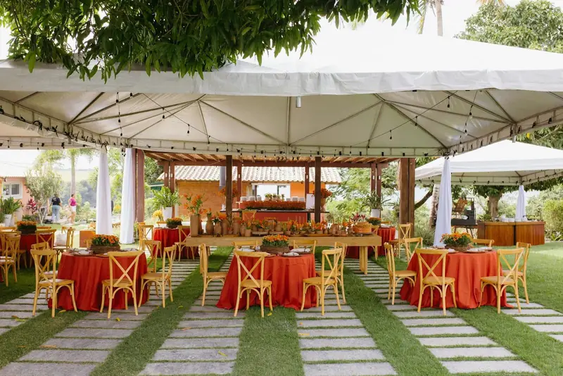 Round tables in orange-red cloths with cross-back chairs under a white canopy on a lawn