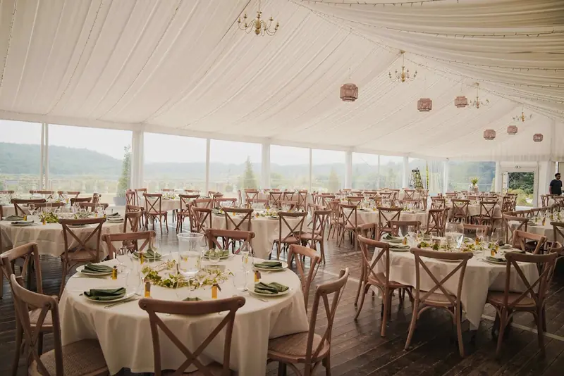 Round tables with cross-back chairs on a wood floor under a draped ceiling with glass walls