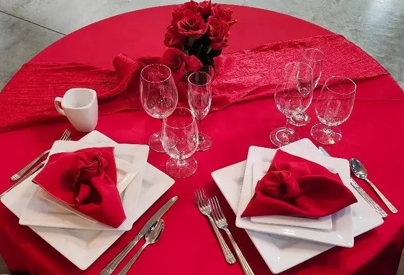 Overhead view of a round table in a red cloth with white plates, red napkins and a vase of red roses