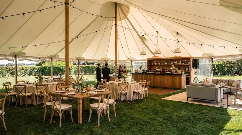 Sailcloth tent over a lawn with wood tables, cross-back chairs, a wood bar and a lounge area