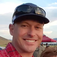 Scott Eastman, managing partner of Celebrate Event Rentals, smiling outdoors in a red plaid shirt and dark cap, with a red event tent and dry hills behind him