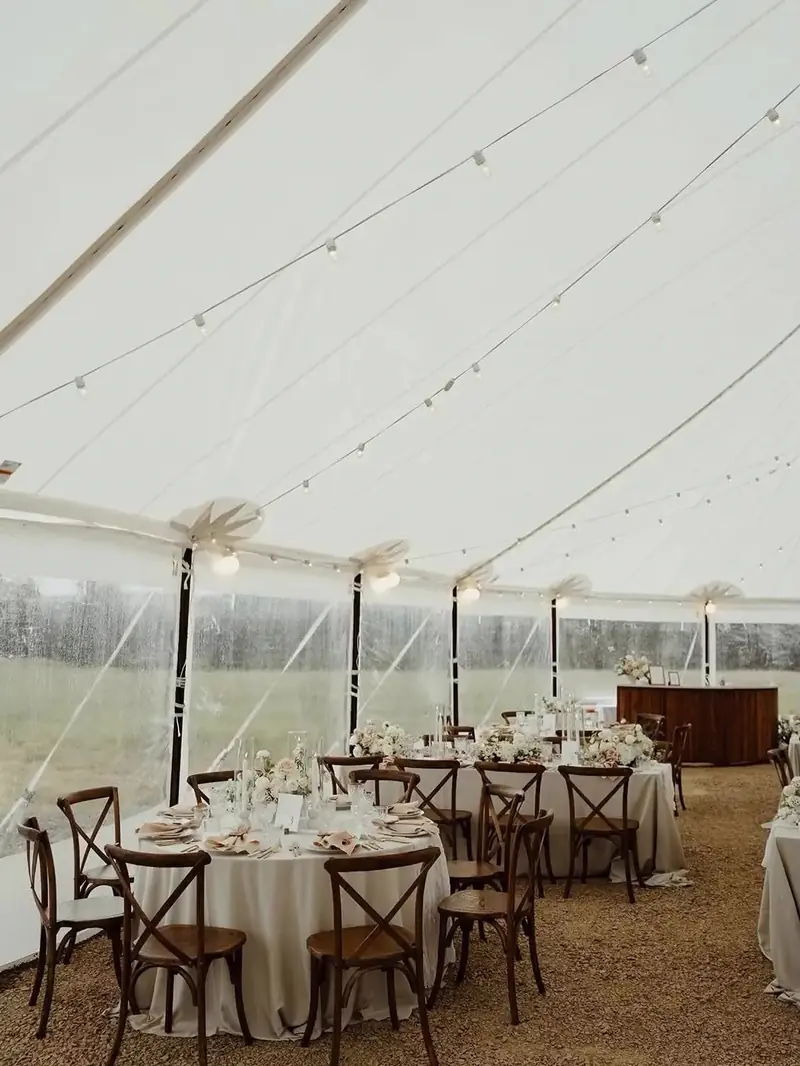 Inside a white tent with clear side walls, round tables in cream cloths, cross-back chairs and a wood bar