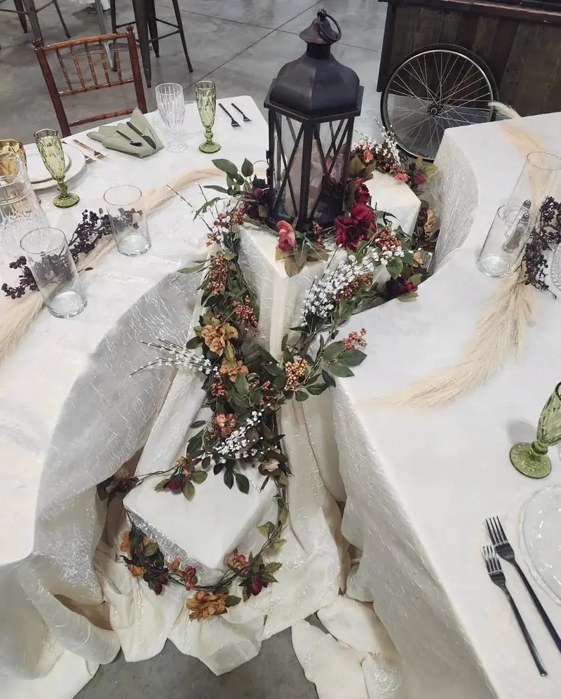 Overhead view of a tiered table display with white cloths, a black lantern and a greenery and berry garland