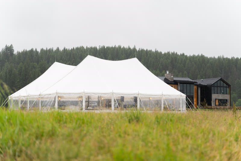 White sailcloth tent with clear sidewalls in a grass field beside a dark wood lodge, tent by Celebrate Event Rentals, Bigfork Montana