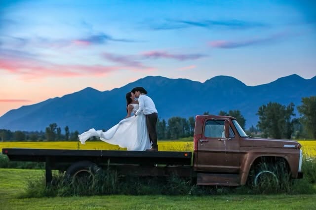 Couple kissing on the bed of a vintage red flatbed truck in a canola field at dusk, mountains behind