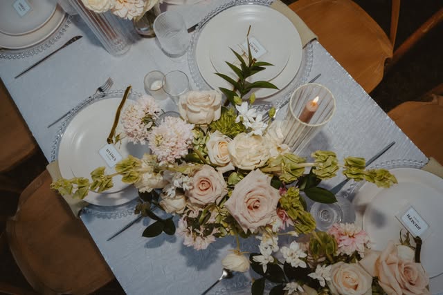 Overhead place setting with blush and cream roses, candles and glass chargers on gray linen