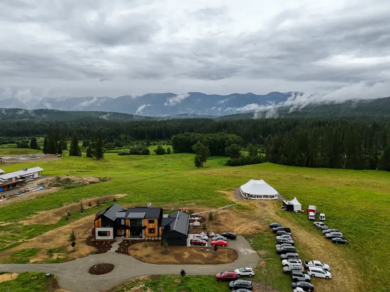 Aerial view of a modern house, parked cars and a white tent on a green field in front of misty mountains