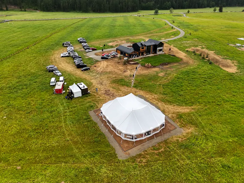 Aerial view of a large white tent beside a modern house and parked cars in a mountain valley