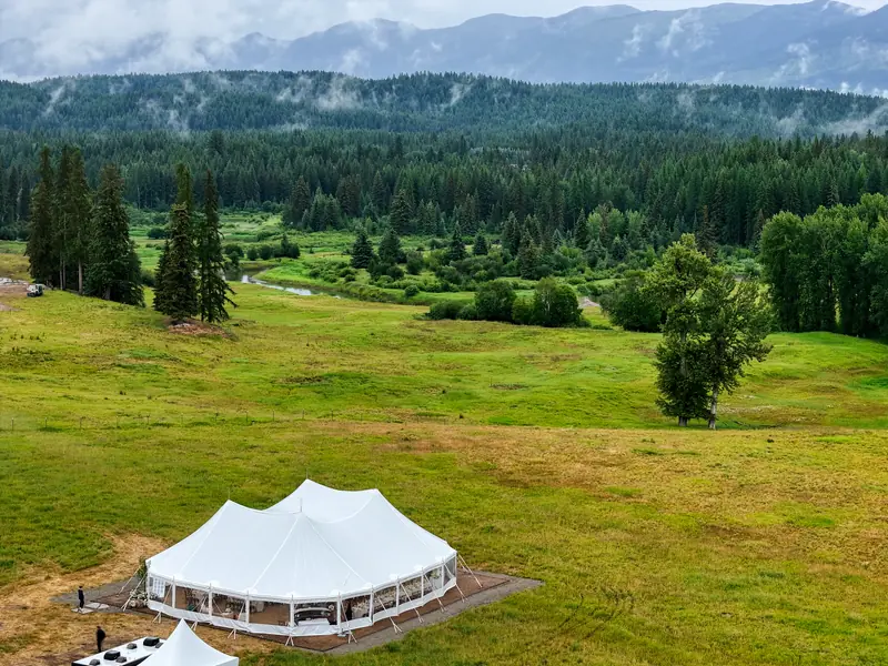 Aerial view of a white event tent pitched in a wide green meadow below forested mountains
