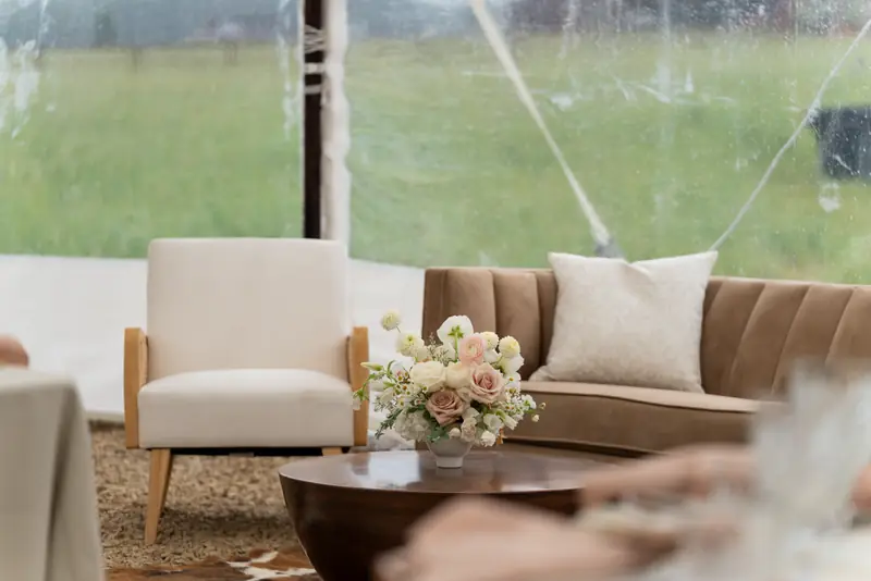 A cream armchair and a brown sofa behind a round wooden coffee table holding a vase of flowers