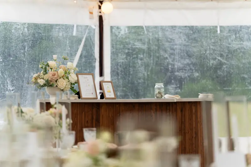 A dark wood bar under a white tent holding a floral arrangement, two framed signs and a glass jar