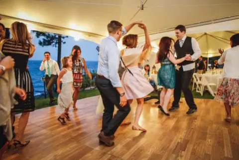Guests dance on a wood plank dance floor inside a white tent near the water
