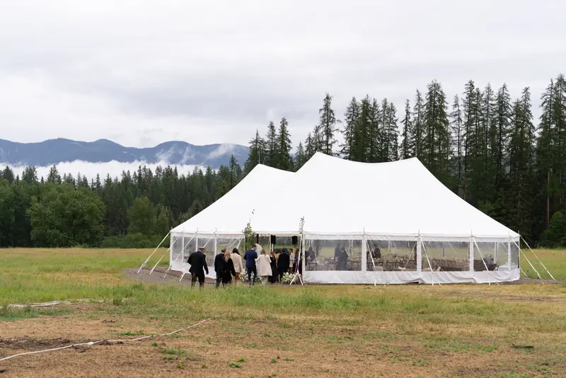 Guests walk across a field toward a white sailcloth tent with pines and misty peaks behind