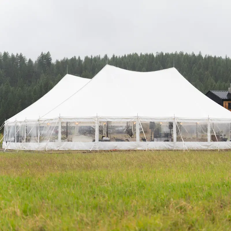 A peaked white tent with clear walls and warm lights inside, set in a grass field beside a modern dark house