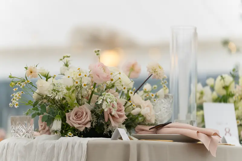 Pink roses and white flowers on a beige tablecloth beside a folded blush napkin and glassware