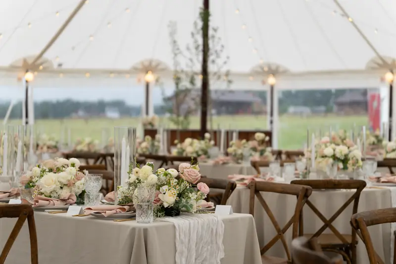 Tables set with beige tablecloths, floral centerpieces, tall glass candles and wooden cross-back chairs inside an illuminated tent
