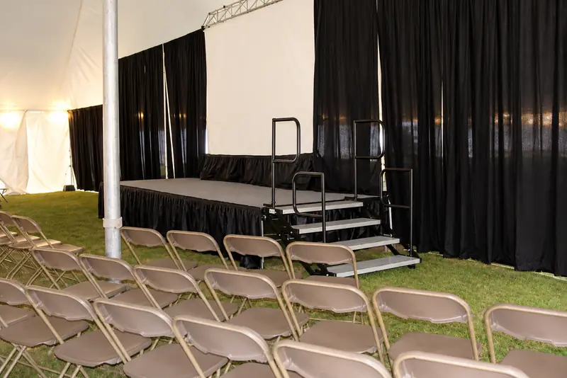 A low stage with a black skirt and metal stairs at one end of a tent, facing rows of folding chairs