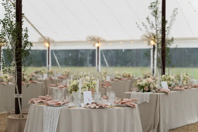 Decorated tables with beige tablecloths, floral centerpieces, crystal glasses and blush napkins inside a large white tent