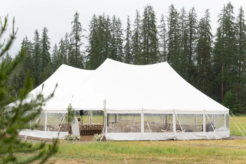 A white tent with three tall peaks and clear walls, with long tables and wood chairs set inside, in front of tall pines