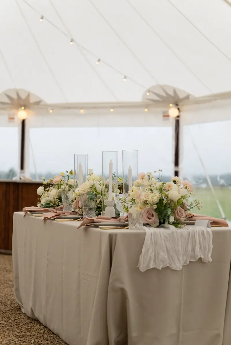A decorated reception table set beneath a white tent hung with string lights, shot in portrait