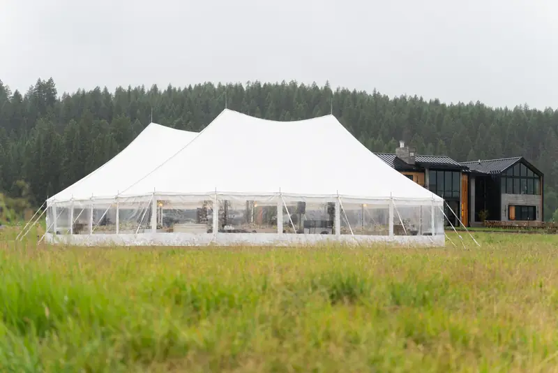 A large white event tent in a grassy field near a modern building with a pine forest in the background