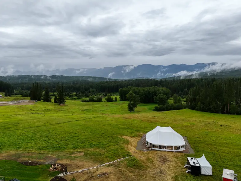 Aerial view of a large white event tent in a green field, backed by evergreen forest and misty mountains