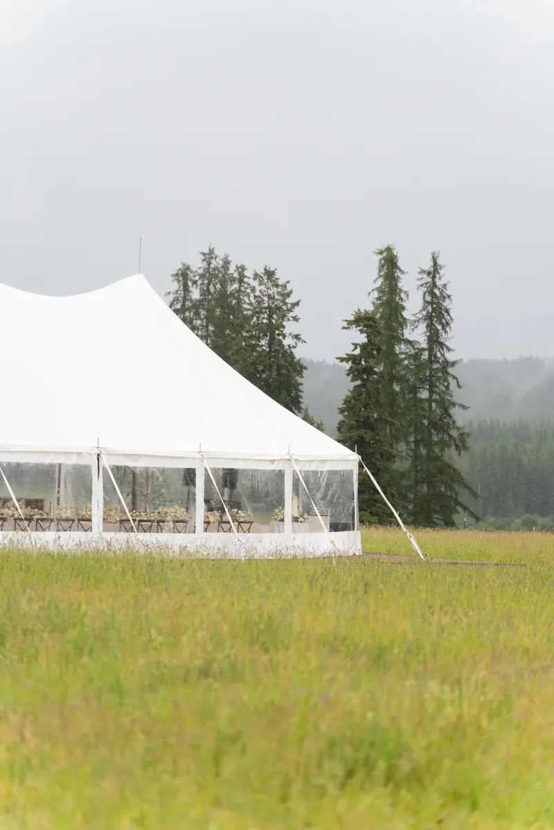 A white event tent set up on a grassy field with evergreen pine trees behind it, shot in portrait
