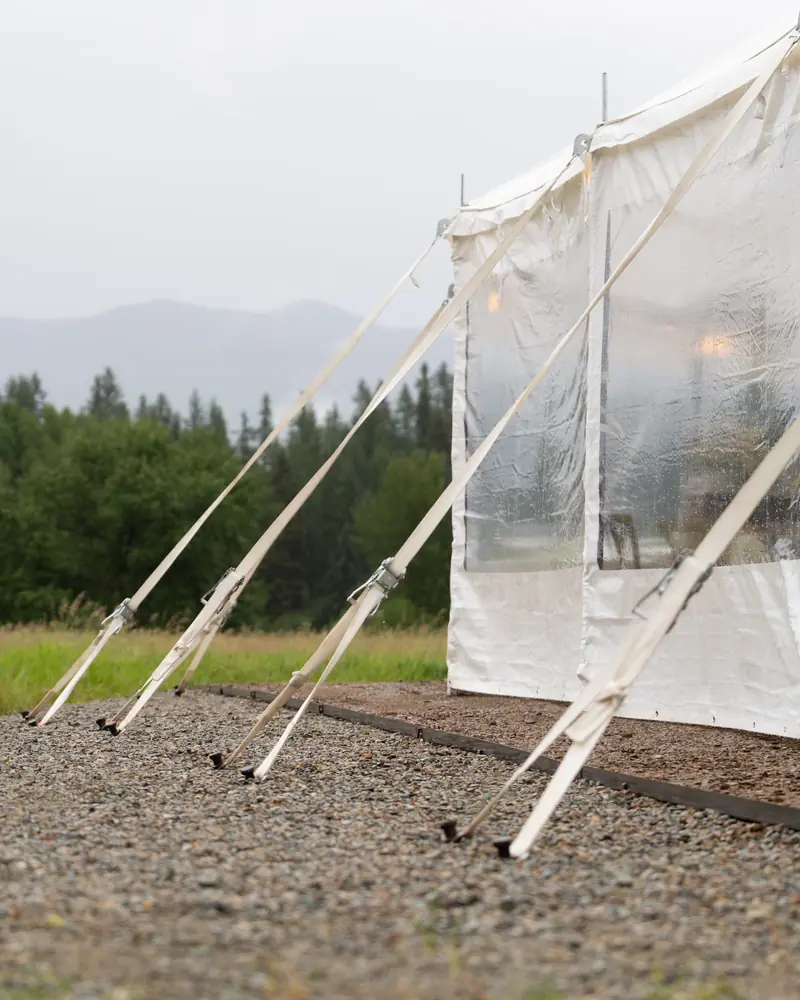 Side of a white tent with clear walls, its guy straps staked into gravel, with forest and misty hills beyond