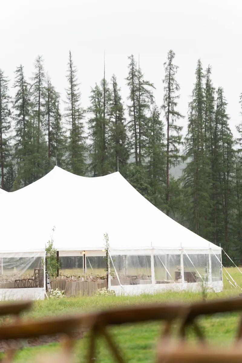 A large white event tent with clear sidewalls in a grassy field against a backdrop of tall pine trees