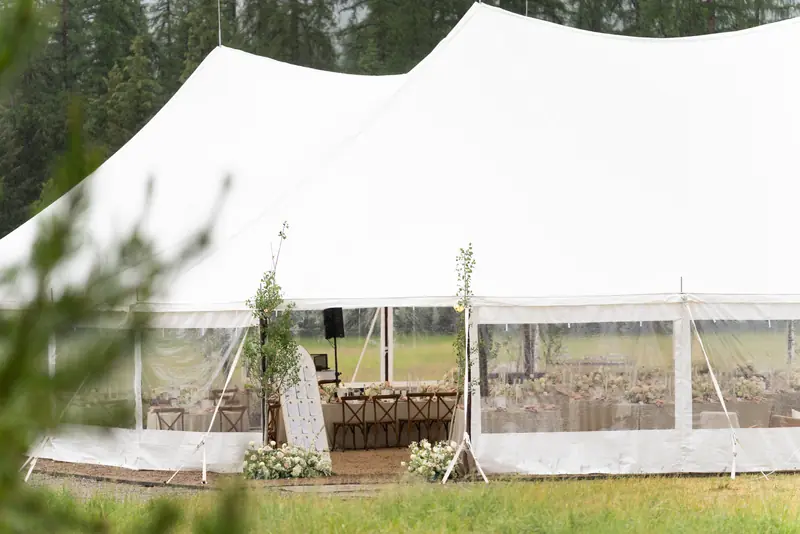 White tent with clear walls and set tables, seen past green trees