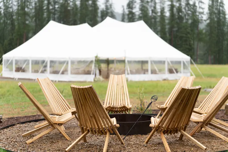Wooden slatted chairs circle a fire pit on gravel, with a large white event tent in the background