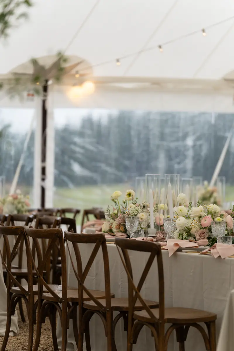 Wood cross back chairs line a table set with flowers, candles, glassware and blush pink napkins inside a white tent