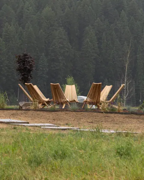 Wooden lounge chairs set in a ring on a lawn with a pine forest behind, under an overcast sky